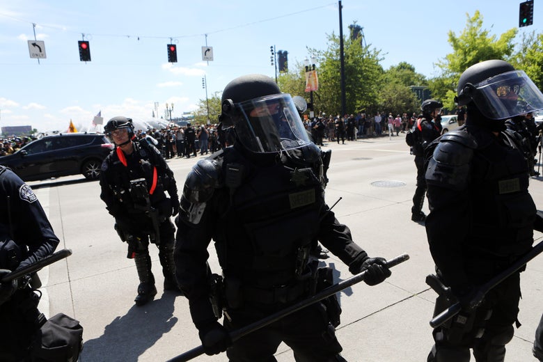 Police in riot gear stand at an intersection. On the other side of the intersection, crowds of protesters can be seen.